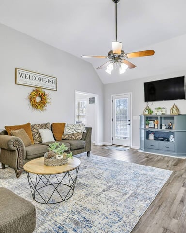 Bright open-plan living room with gray upholstered seating, wood coffee table, ceiling fan, wall-mounted TV, and blue storage cabinet