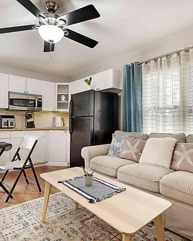 Open-plan living room with beige sofa, dining table, kitchen with white cabinets and black refrigerator, ceiling fan, and large windows with sheer curtains.