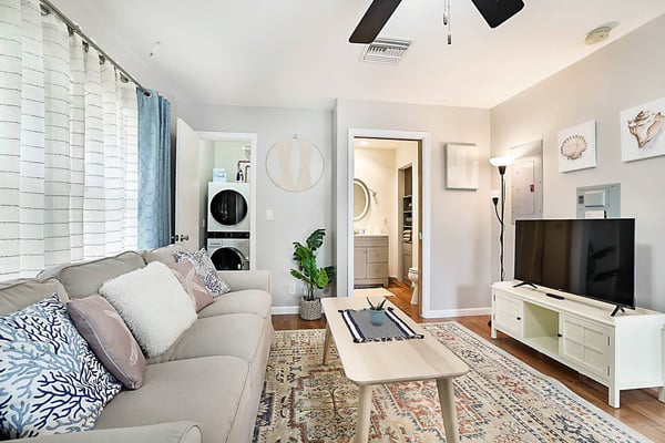 Open-plan studio with gray sofa, TV on white console, stacked washer-dryer, bathroom and kitchenette visible through doorways