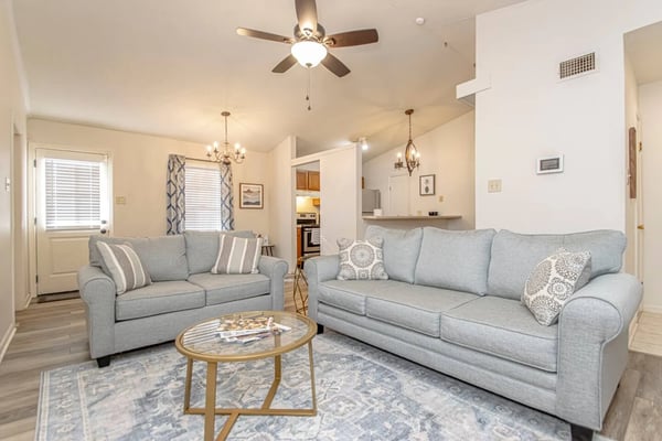 Open-plan living room with two gray sofas, round glass table, ceiling fan, and kitchen visible in background