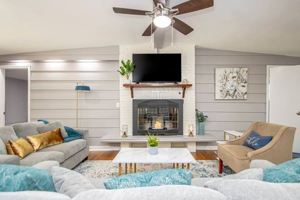 Living room with fireplace, TV on mantel, ceiling fan, gray sofa with gold pillows, tan armchair, and marble coffee table.