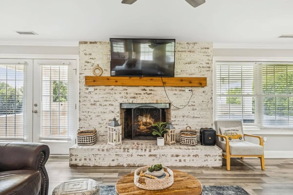 Living room with whitewashed brick fireplace, mounted TV on wooden mantel, and wood-framed armchair
