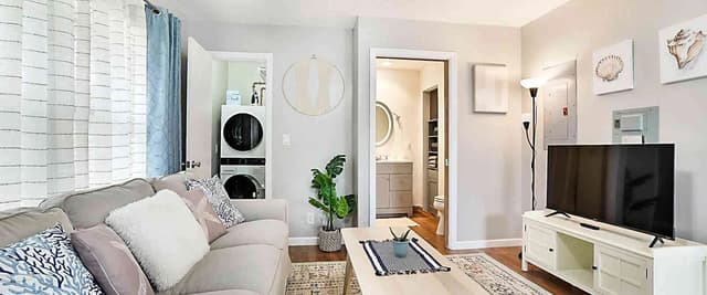 Studio apartment with gray sofa, TV on white media console, coffee table, and open bathroom and laundry area visible through doorways