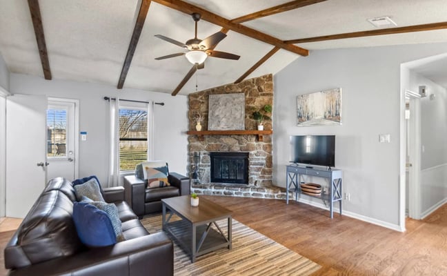Open-plan living room with exposed wood beams, stone fireplace, leather seating, and TV entertainment center