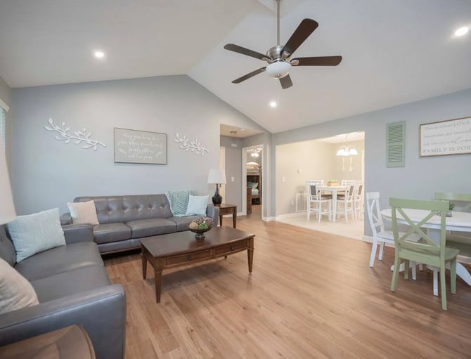 Open-plan living room with gray sectional sofa, wood coffee table, ceiling fan, and dining area with green table visible in background.